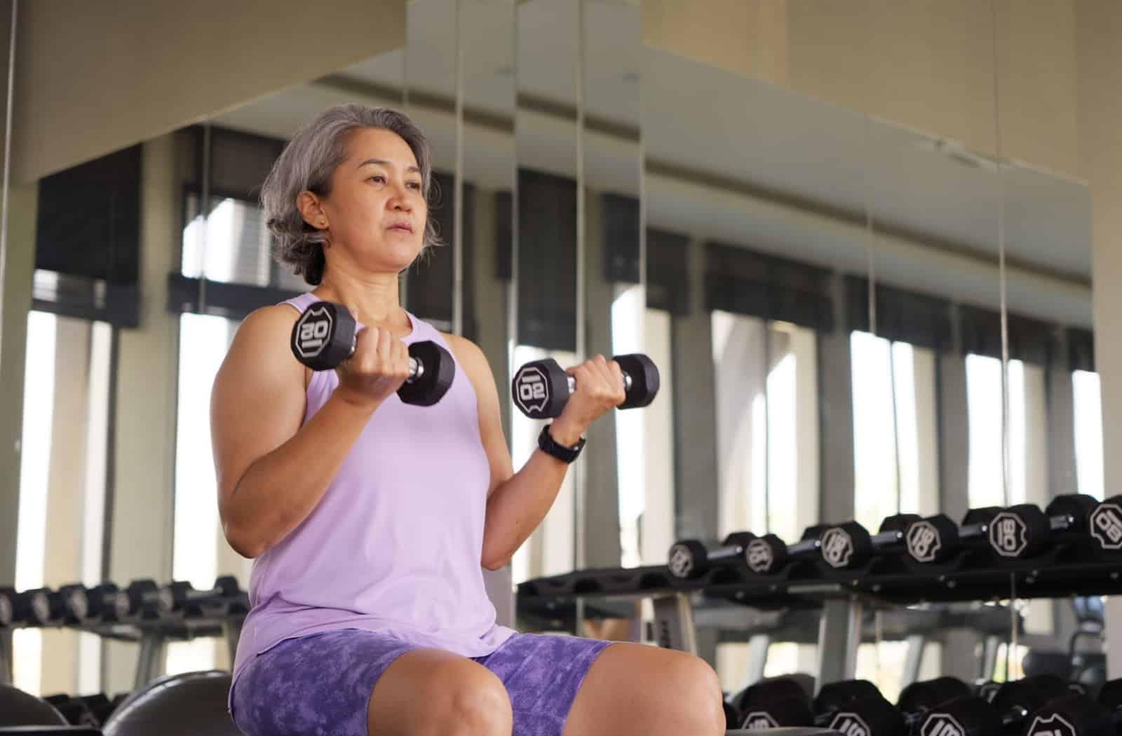 A woman sitting on a workout bench, holding two dumbbell weights during her resistance exercise routine to help manage perimenopause fatigue.
