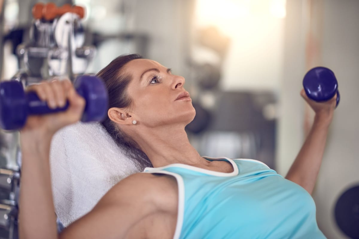 Woman performing dumbbell chest press exercise on bench in gym wearing blue workout top.